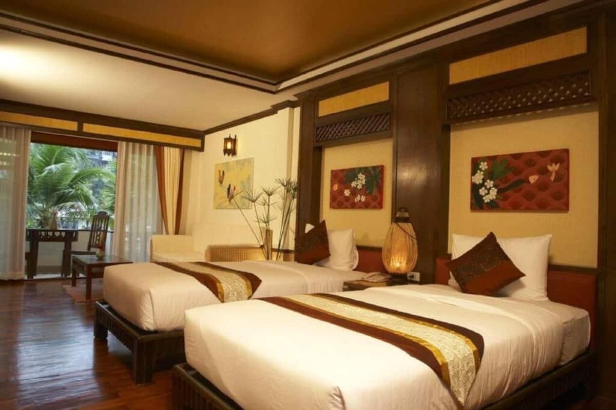 Quarto de hotel com duas camas, um espaço de estar com mesa e cadeiras, e vista para palmeiras pela janela.