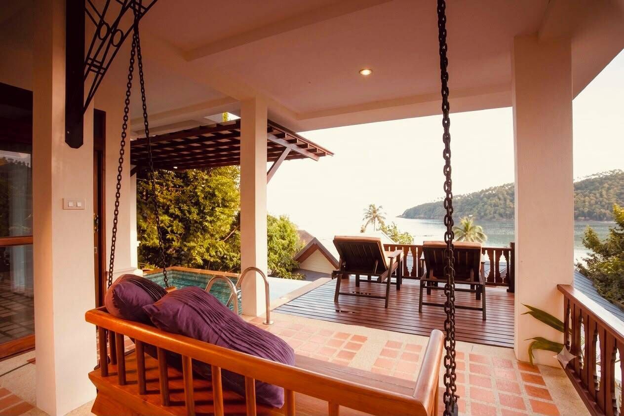 Una silla mecedora de madera con cojines, una mesa de madera con sillas, y vista al mar y colinas.