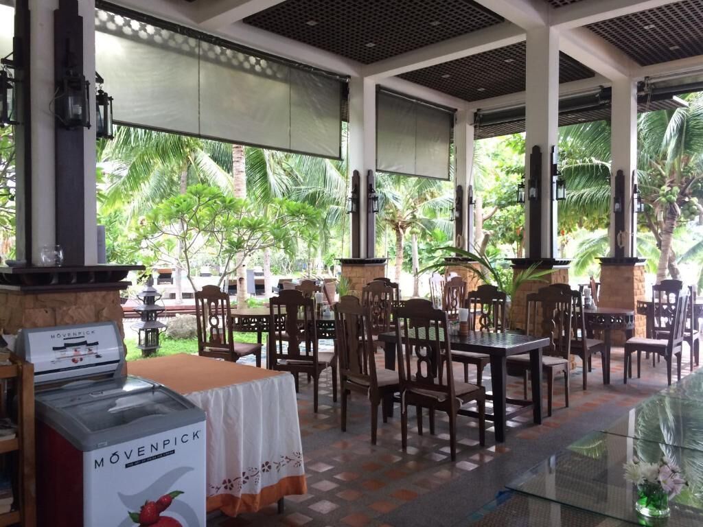 A covered outdoor dining area with tables and chairs, a movable ice cream machine, and a view of palm trees.