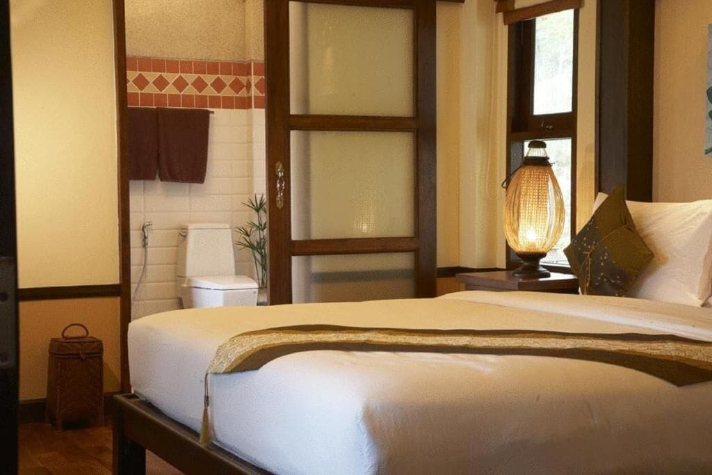Habitación de hotel con cama, mesita de noche con lámpara, ventana con cortinas y baño visible a través de una puerta abierta.
