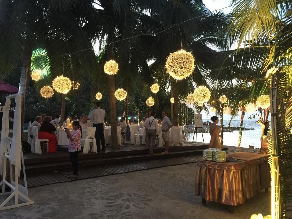 An outdoor event with round, illuminated decorations hanging from the ceiling, surrounded by palm trees and a stage with tables set for an event.