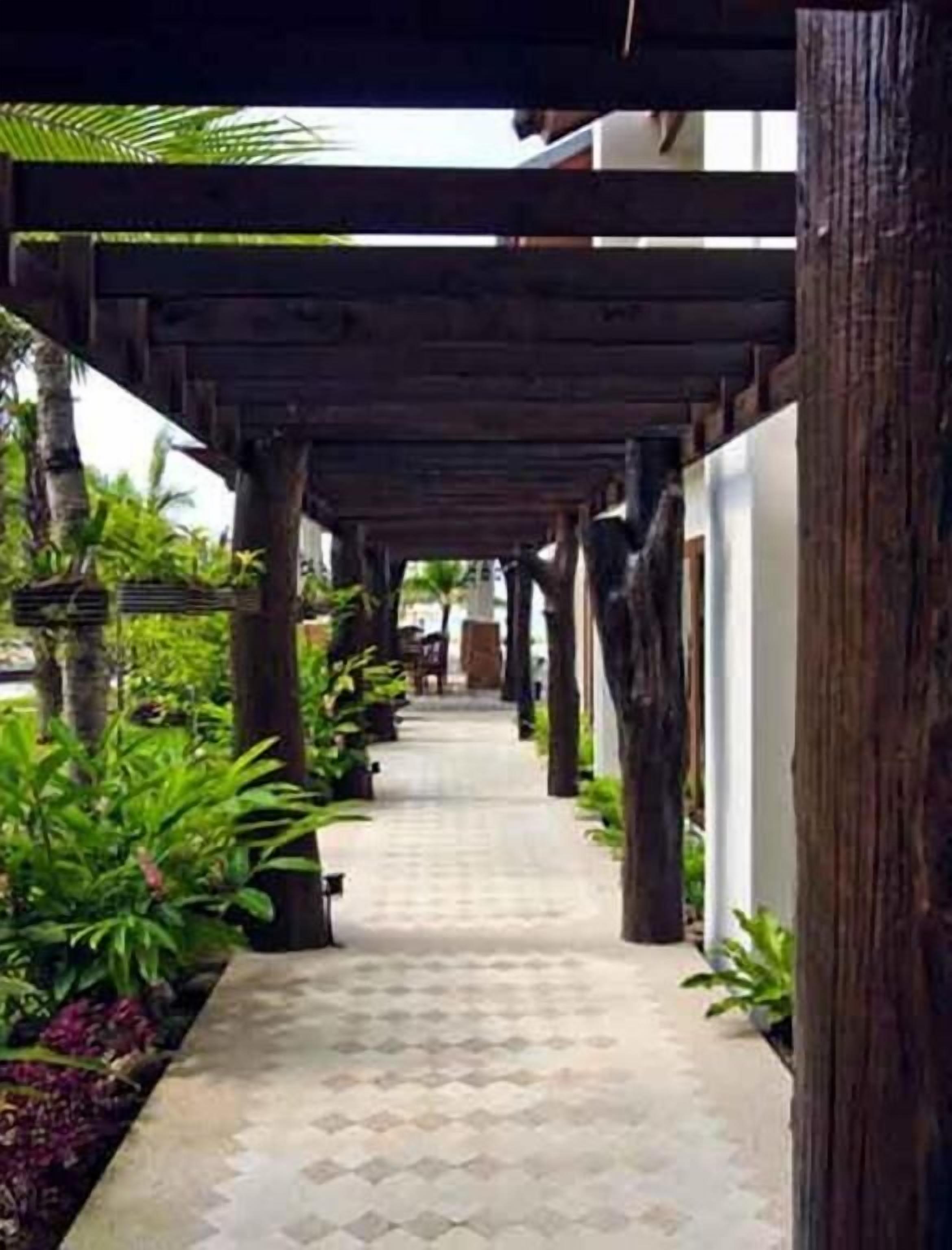 A covered walkway with wooden pillars, surrounded by greenery and a tiled floor.
