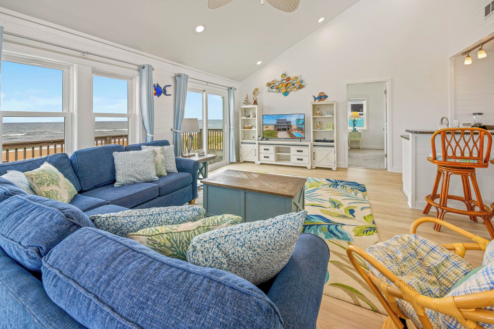 A spacious living room with a large sofa, a coffee table, and a wicker chair. There is a TV on a white cabinet, and a view of the ocean through large windows.