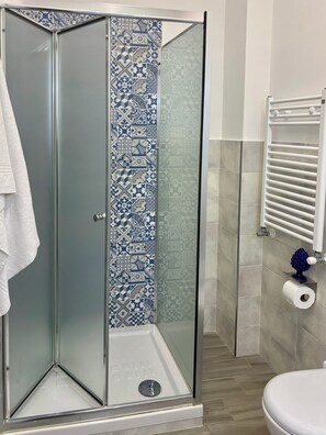 Una doccia con porta in vetro, un asciugamano bianco appeso al muro e un water con dispenser di carta.