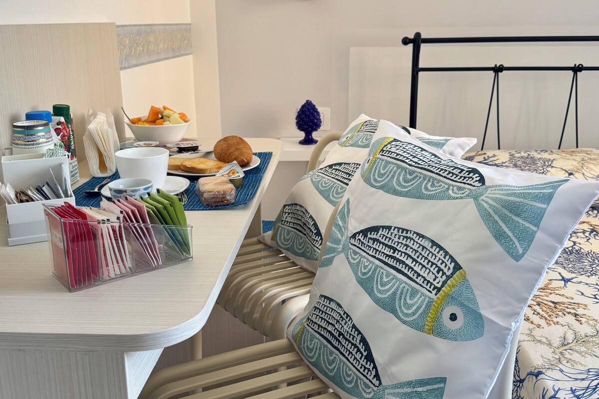 A neatly arranged hotel room with a bed, a bedside table with a bowl of fruit, a chair, and a desk with various items.