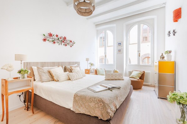 Een ruime slaapkamer met een groot bed, een houten nachtkastje, een bijzettafeltje met een lamp en uitzicht op de stad door grote ramen.