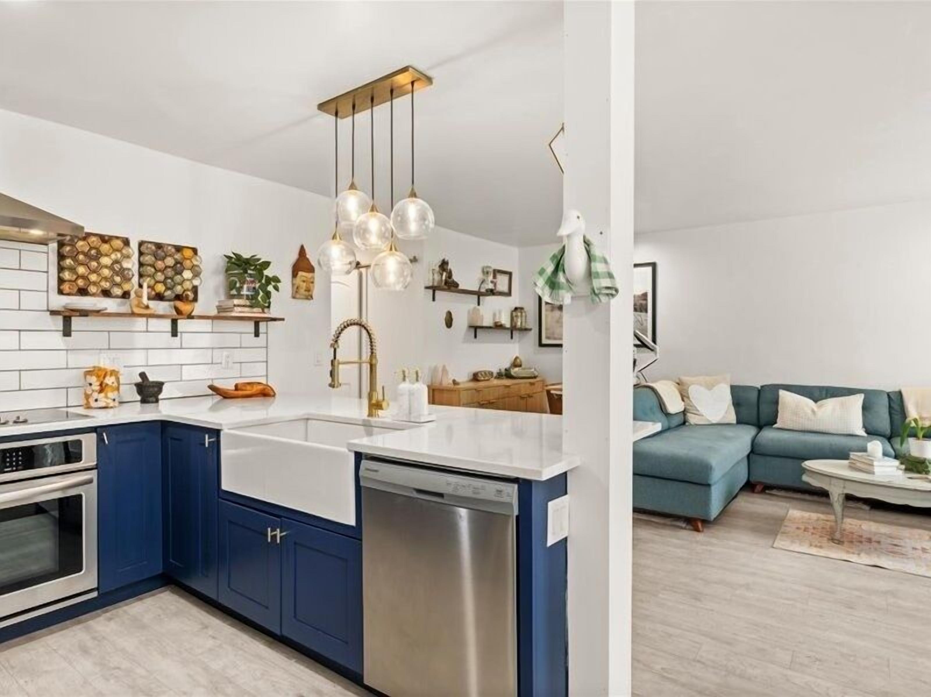 A modern kitchen with blue cabinets, a white countertop, and a stainless steel dishwasher. There is a hanging light fixture with clear glass shades.