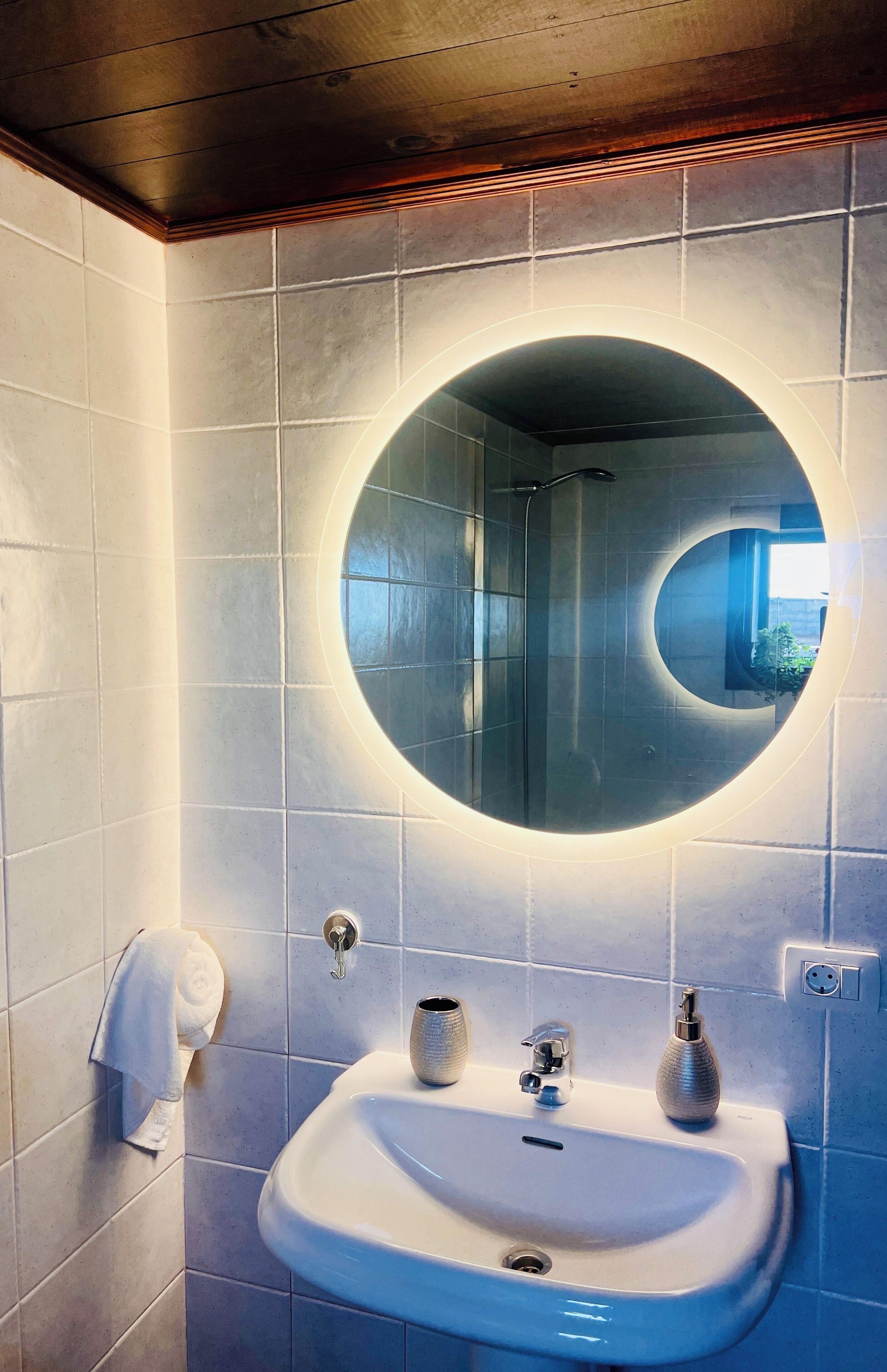 A bathroom with a round mirror, a sink, a towel dispenser, and a shower head.