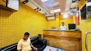 A room with a patterned yellow wall, a wooden reception desk, a black leather couch, and a ceiling fan. - Hotel zam Zam Residential (Dhaka)