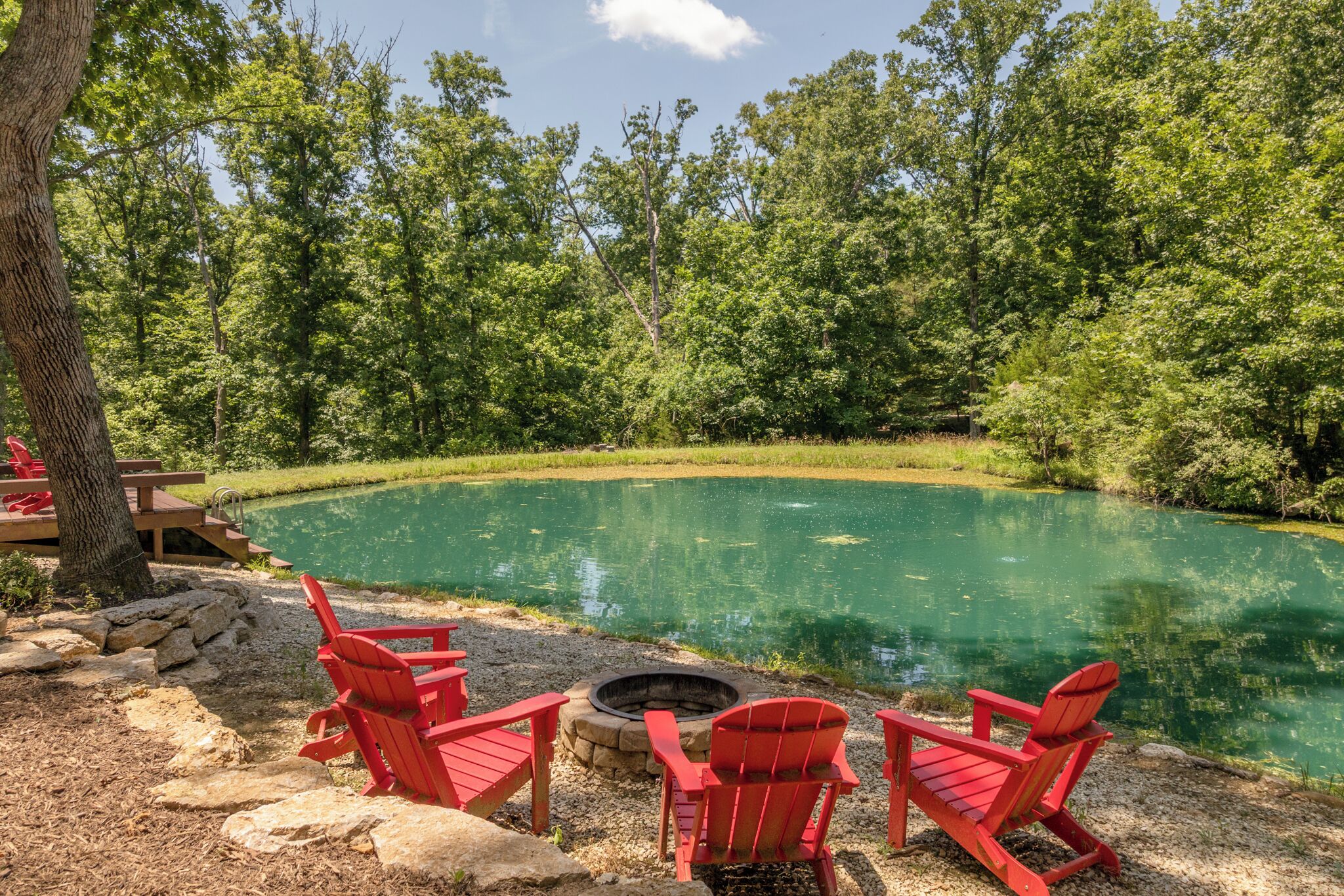 A serene outdoor setting with a circular pond, surrounded by dense greenery and red Adirondack chairs.