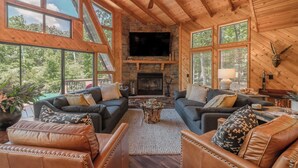 A spacious log cabin interior with a fireplace, leather sofas, and large windows offering a view of the forest.