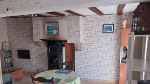 A rustic room with exposed wooden beams, a stone wall, a fireplace, a dining table with a tablecloth, and a bookshelf.