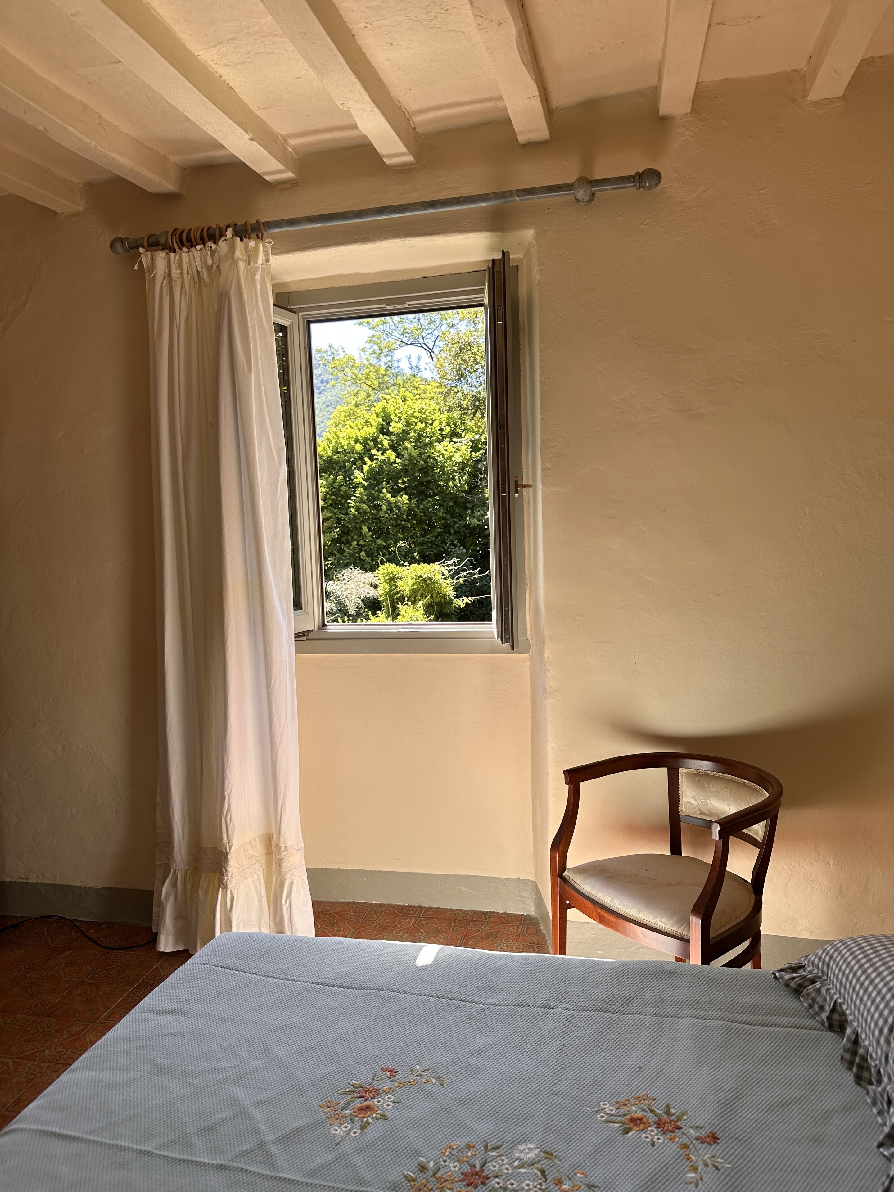 Een slaapkamer met een bed, een stoel, een raam met gordijnen en uitzicht op groen.