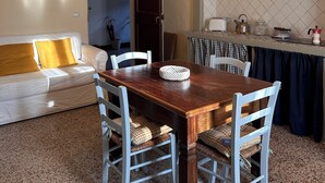 Un coin repas avec une table en bois et des chaises blanches, un canapé blanc avec des coussins jaunes, et un espace cuisine avec un dosseret carrelé et une cuisinière.