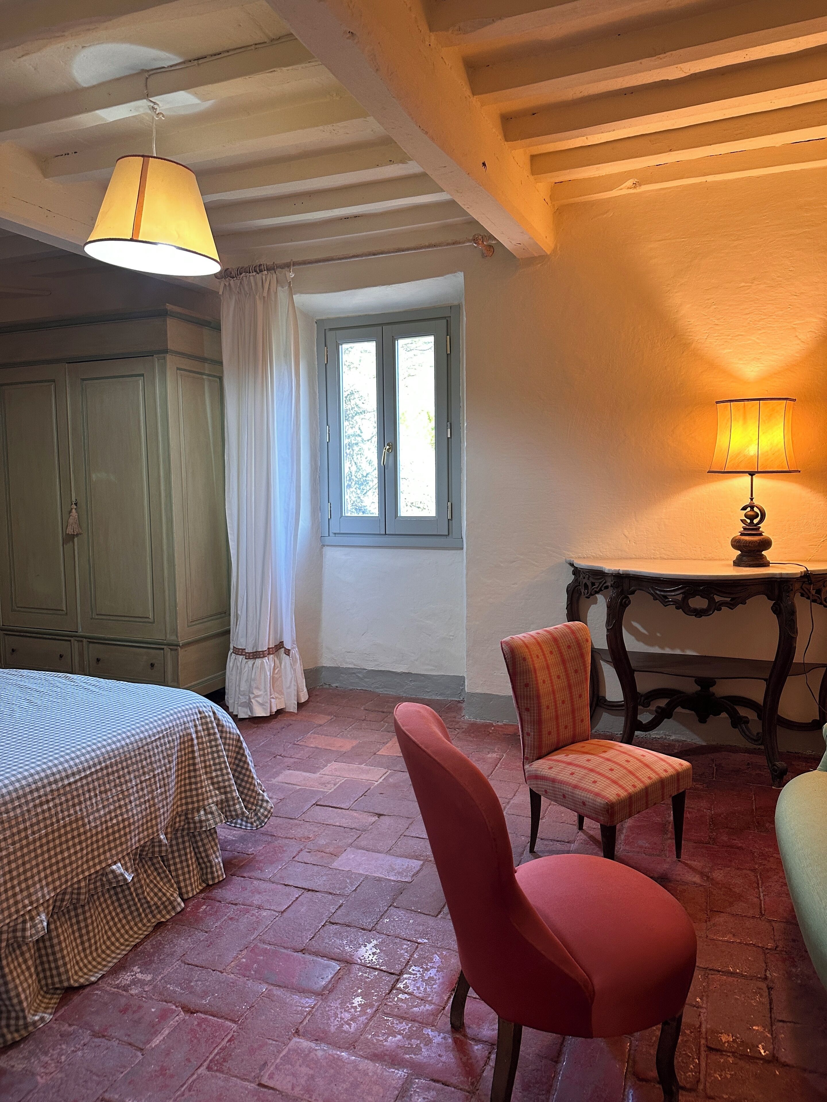 Una habitación con edredón a cuadros, una silla roja, una mesa con lámpara y una ventana con cortinas.