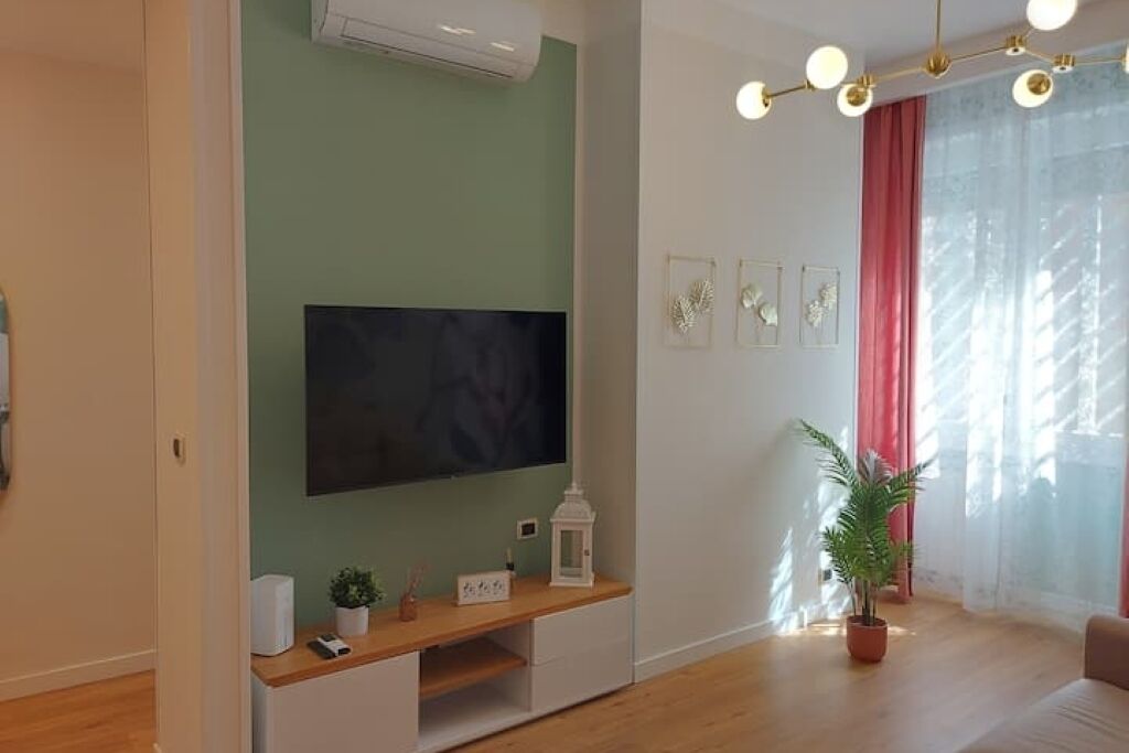 A living room with a flat-screen TV mounted on a green wall, a wooden entertainment unit below, a potted plant, and a red curtain.