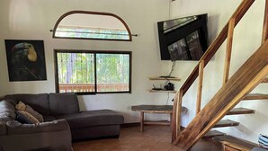 Un salon comprenant un canapé, un fauteuil, un téléviseur, une fenêtre donnant sur un paysage verdoyant et un escalier en bois.