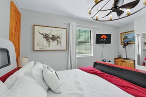 A bedroom with a bed, a TV, a painting, a window with blinds, a ceiling fan, and a dresser.