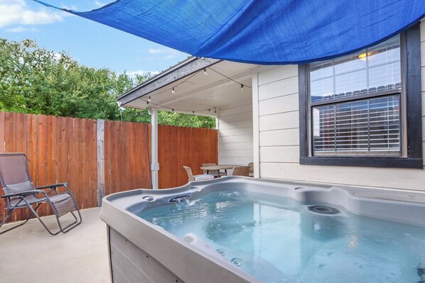 A hot tub in a backyard with a wooden fence, a chair, and a table under a blue shade.
