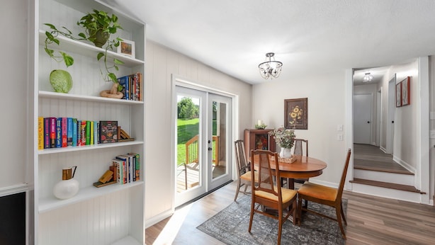 Uma sala de jantar com mesa e cadeiras de madeira, uma estante com livros e objetos decorativos, um lustre e vista para o exterior por uma porta de vidro deslizante.