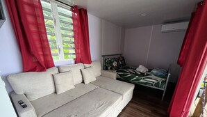 A room with a beige sofa, a bed with a tropical print, red curtains, and a wooden floor.