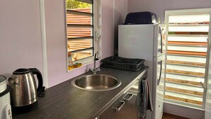 A compact kitchen with a stainless steel sink, a kettle, a refrigerator, and a window with blinds.