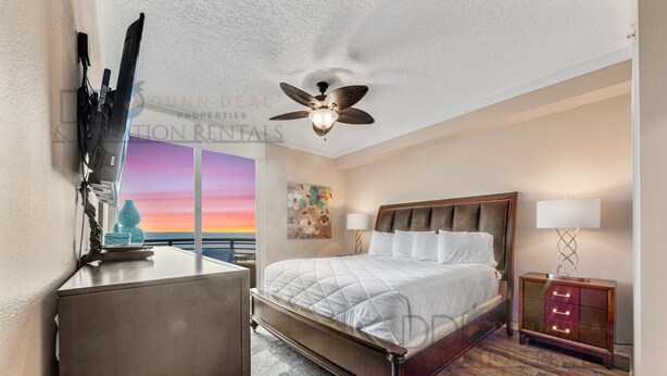 A bedroom with a bed, a ceiling fan, a nightstand, and a view of the sunset through the window.