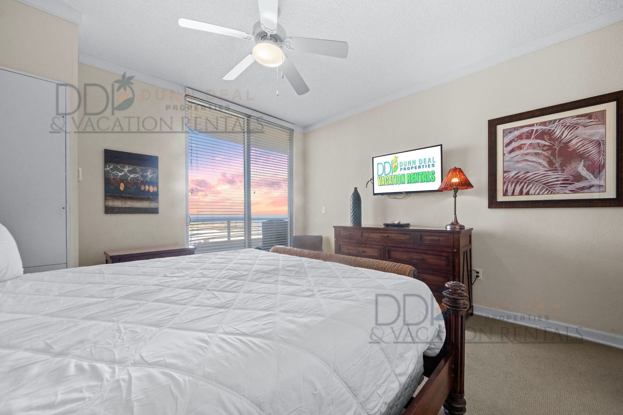 Ein Schlafzimmer mit einem großen Bett, einem Deckenventilator, einem Fernseher, einem Nachttisch und einem Fenster mit Sonnenuntergangsblick.