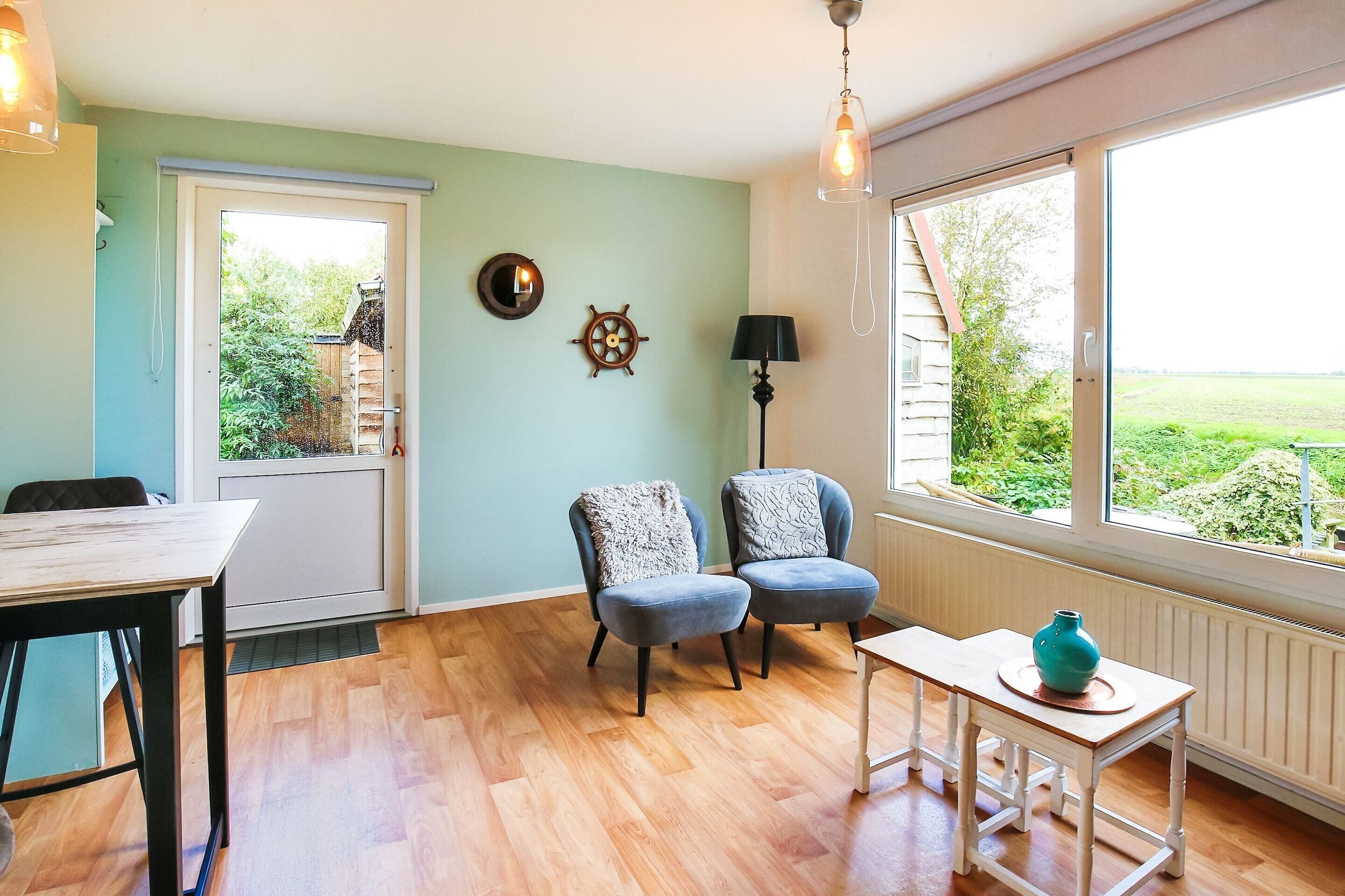Een kamer met een houten vloer, een witte tafel, twee blauwe stoelen, een lamp, een wandklok en uitzicht op groen buiten.