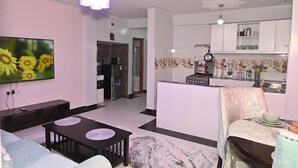 Un salon comprenant un téléviseur, un canapé, une table basse et un coin cuisine avec une cuisinière et des placards.