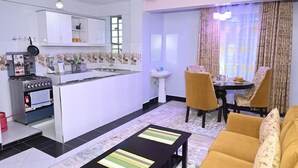 Une cuisine moderne avec des meubles blancs, une cuisinière à gaz et un évier. On y trouve un coin repas avec une table et des chaises, ainsi qu’un salon avec un canapé et une table basse.