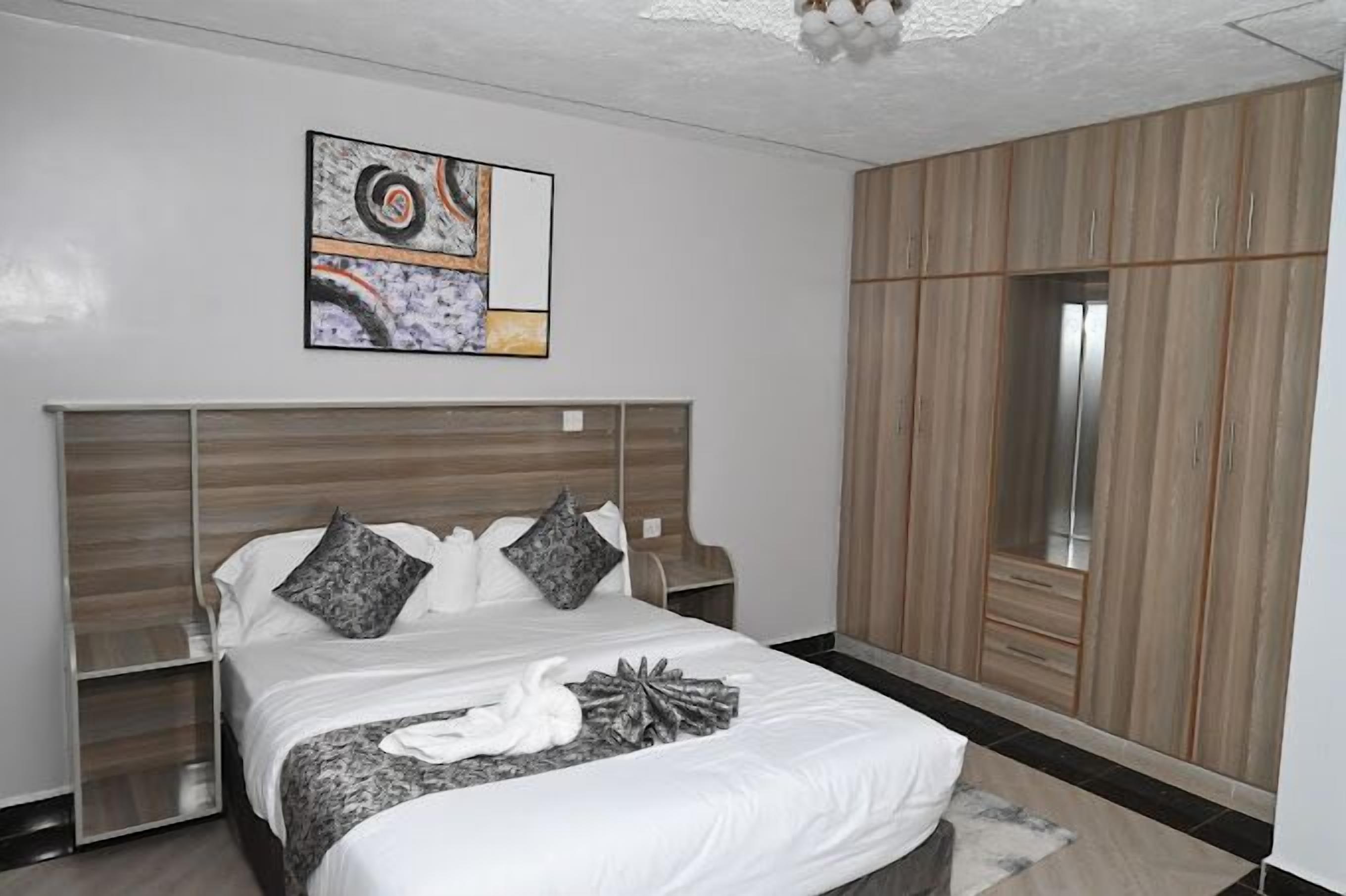 A modern bedroom with a large bed, a headboard with built-in storage, a wardrobe, and a wall-mounted artwork.