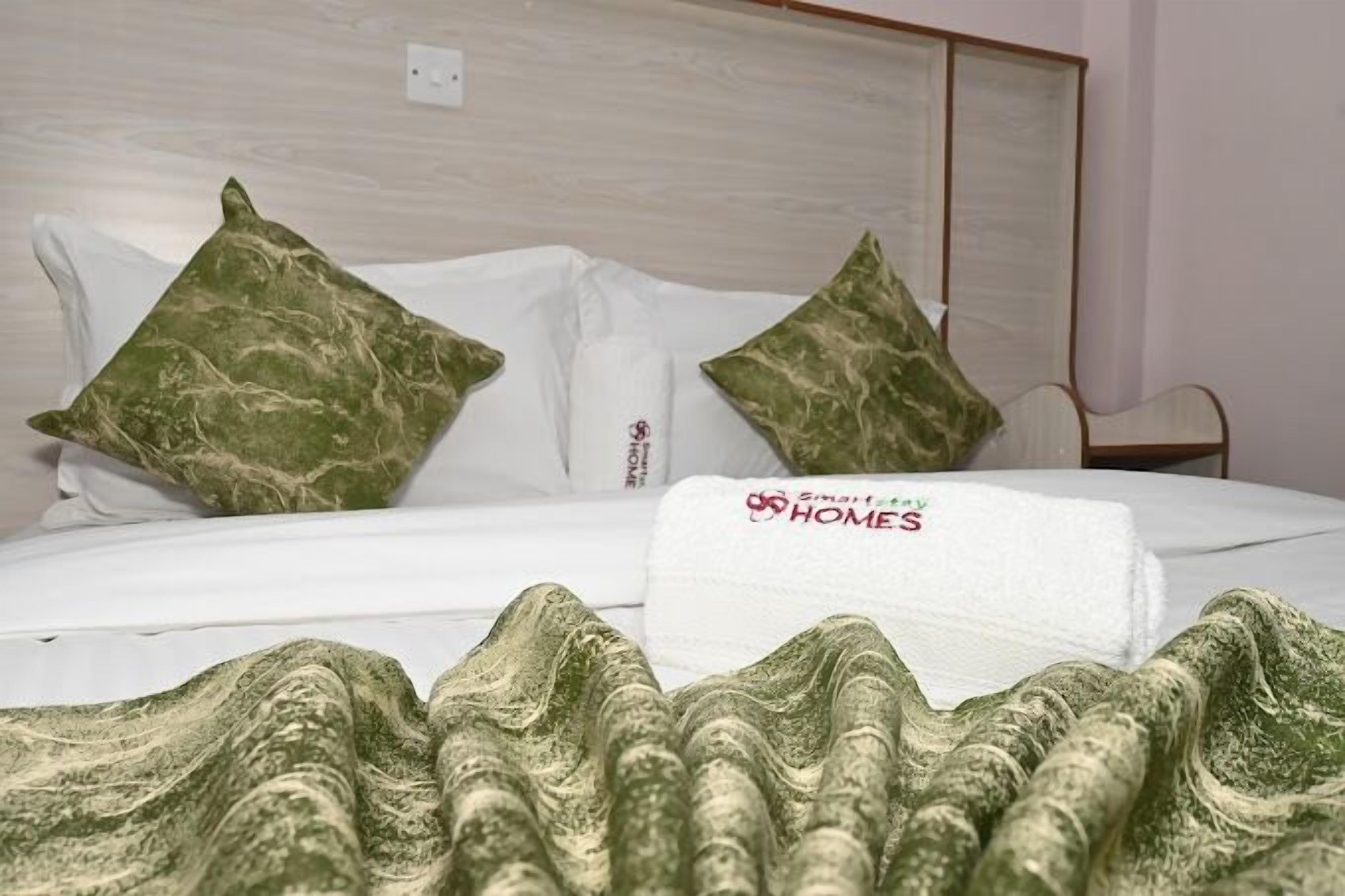 A neatly made bed with a white sheet and two green pillows, a white towel with a logo, and a green patterned blanket.