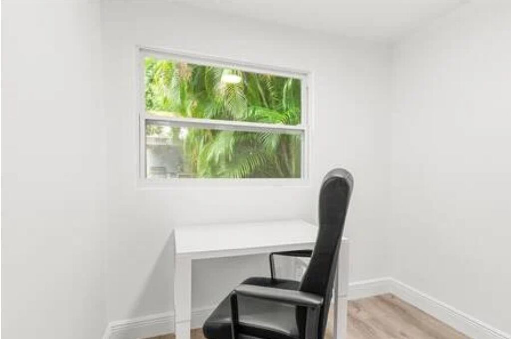 Un escritorio blanco con una silla negra en una habitación con ventana que da a zonas verdes.