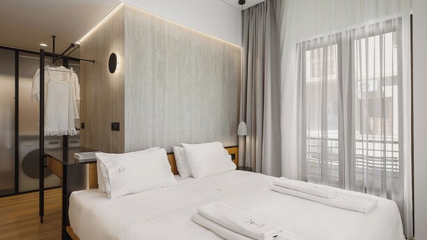 Una habitación de hotel con una cama grande, un armario, un escritorio y un ventanal con cortinas translúcidas.