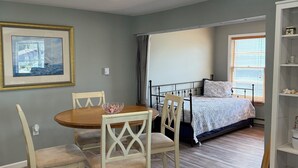 A room with a bed, a dining table with chairs, a framed picture on the wall, and a bookshelf.
