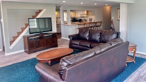 A modern living room with a leather sofa, a wooden coffee table, and a flat-screen TV on a wooden stand. A staircase leads to an upper level.