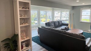 A living room with a black leather sofa, a white shelving unit, and a view of a residential area through large windows.