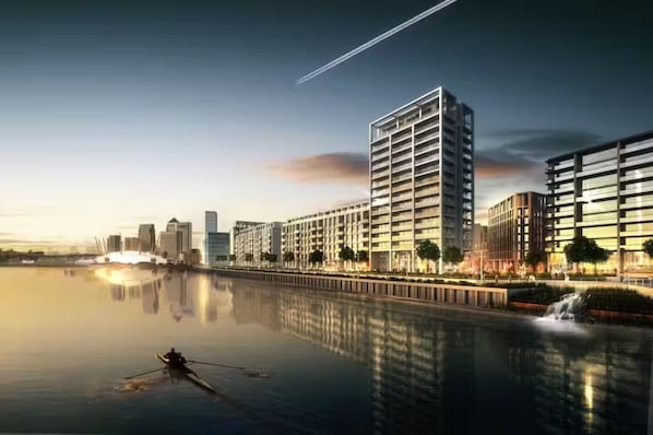 Un edificio moderno de gran altura con su reflejo en aguas tranquilas, rodeado de otros edificios altos y un cielo despejado.