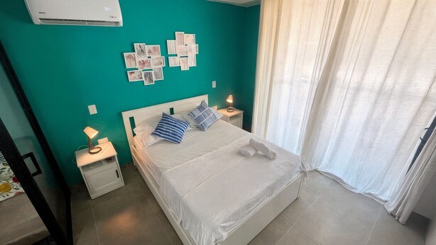 Un dormitorio ordenado con una cama, dos mesitas de noche con lámparas, un aire acondicionado de pared y una ventana con cortinas.