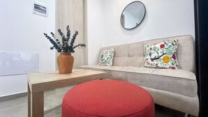Una sala de estar moderna con una mesa de madera, un otoman rojo, un sofá beige con cojines estampados, un espejo redondo y una planta decorativa en un florero tejido.