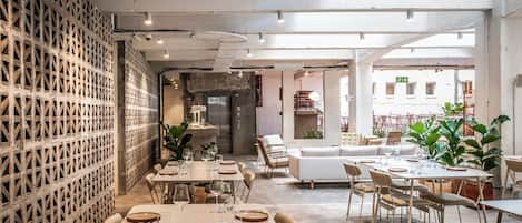 Un comedor interior con mesas dispuestas para dos, sillas y una pared decorativa con diseño geométrico.