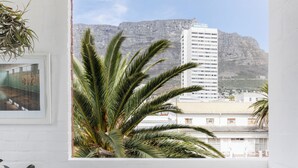 A balcony with a white chair, a small table, and a potted plant. A window offers a view of a building and palm trees. - Blackbrick Hotel Gardens - Cape Town (Cape Town)