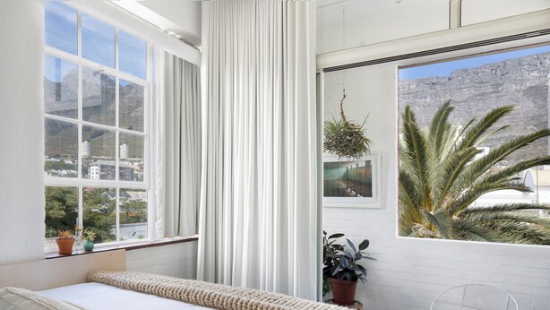 A bedroom with a large window, a bed with a white comforter, a potted plant, and a view of a mountain. - Blackbrick Hotel Gardens - Cape Town (Cape Town)