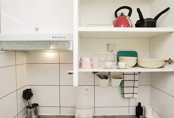 Una cocina con paredes de azulejos blancos, encimera blanca y estantes con utensilios de cocina.