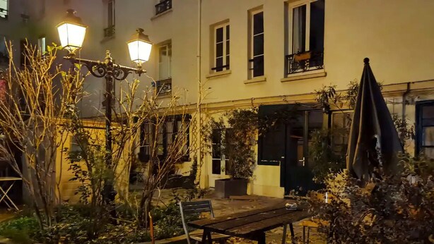 Un patio al aire libre con mesa y sillas de madera, rodeado de plantas y un farol.