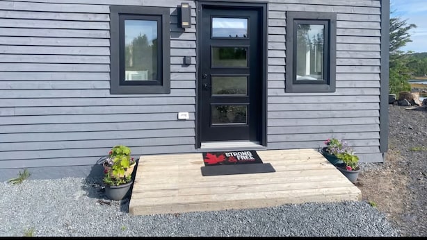A modern house with a grey exterior, a black door, and two windows. There is a wooden deck with a mat that says "STRONG FREE." - The Northern Tickle. 20 minutes to Twillingate. Gorgeous views of the ocean. (Summerford)