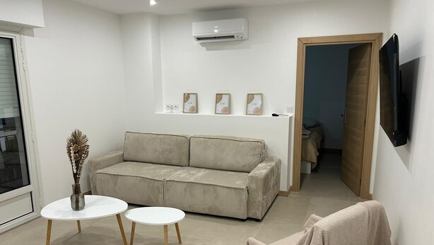 Una sala de estar moderna con un sofá color beige, dos mesas redondas blancas, un jarrón decorativo y un televisor montado en la pared.