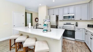 Uma cozinha moderna com armários brancos, uma ilha central com banquetas, forno embutido, micro-ondas e liquidificador.