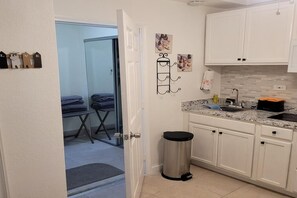 A compact kitchen with white cabinets, a stainless steel trash bin, and a countertop with a sink and various items. - Charming 1-bedroom house in terrific Delray Beach with AC (Delray Beach)